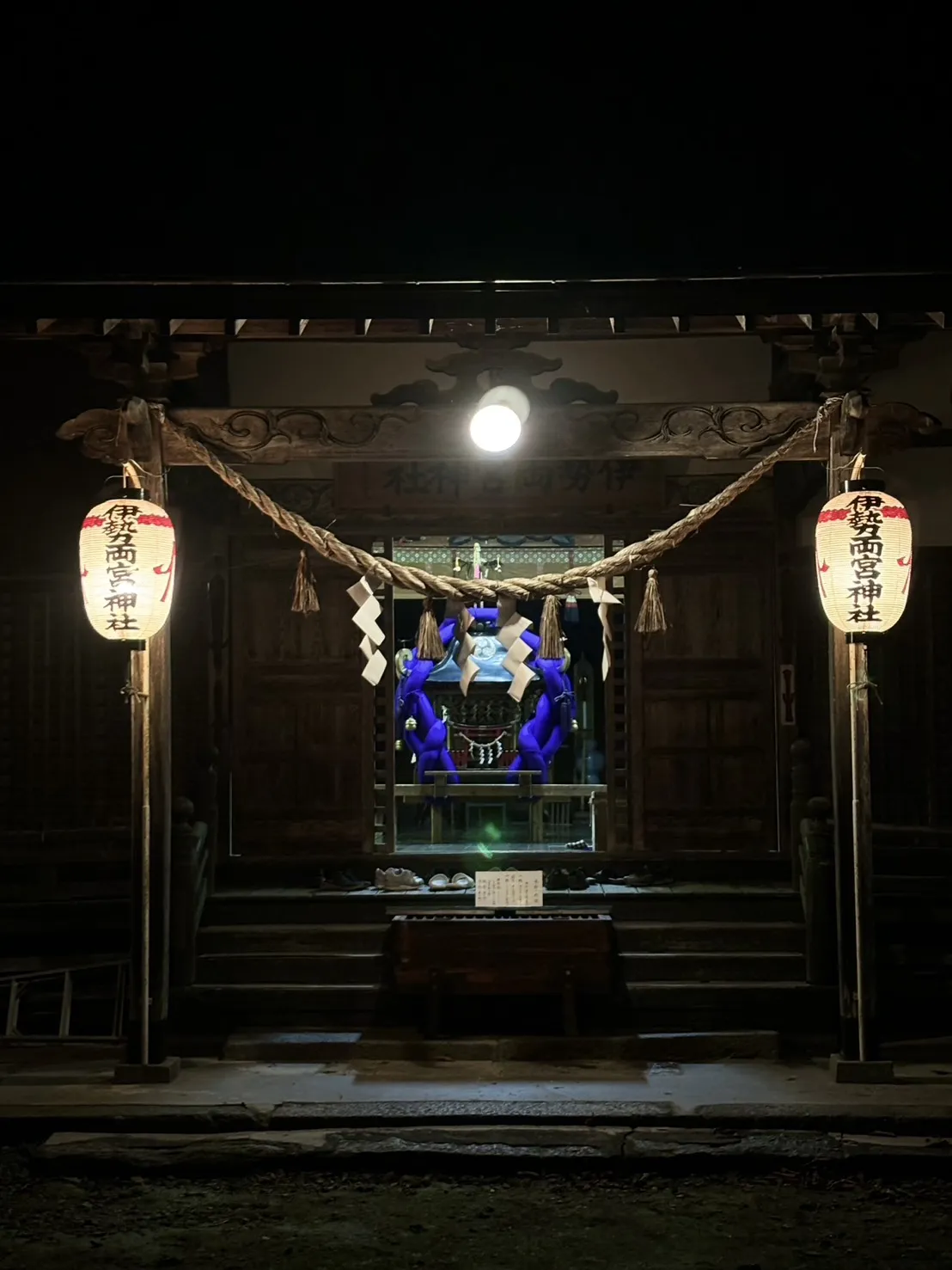 夜の伊勢両宮神社拝殿。両側に「伊勢両宮神社」と記された赤提灯が灯り、注連縄と御輿が見える。